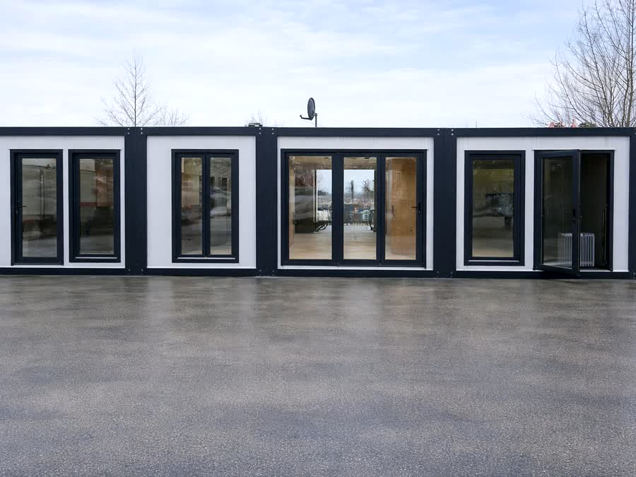 Vier verbundene Container-Module mit anthrazitfarbenem Rahmen und bodentiefen Fenstern auf einem Hof